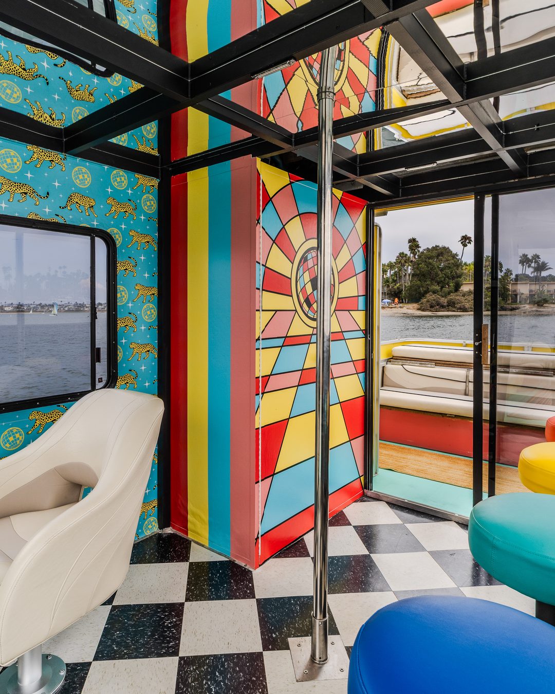 Disco Boat interior — rainbow-stripe wall, chrome dance pole, jewel-tone stools on the checkerboard floor